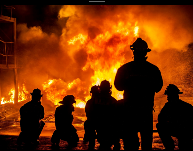 Silhouettes of firefighters battling a large blaze at night, highlighting the intense flames and smoke that fill the scene.