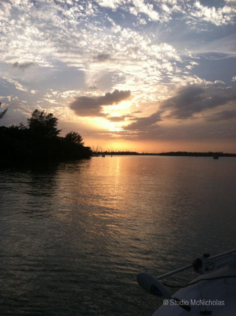 Sunset over a calm waterway, reflecting vibrant orange and purple hues. Silhouetted trees line the shore, enhancing the tranquil atmosphere.