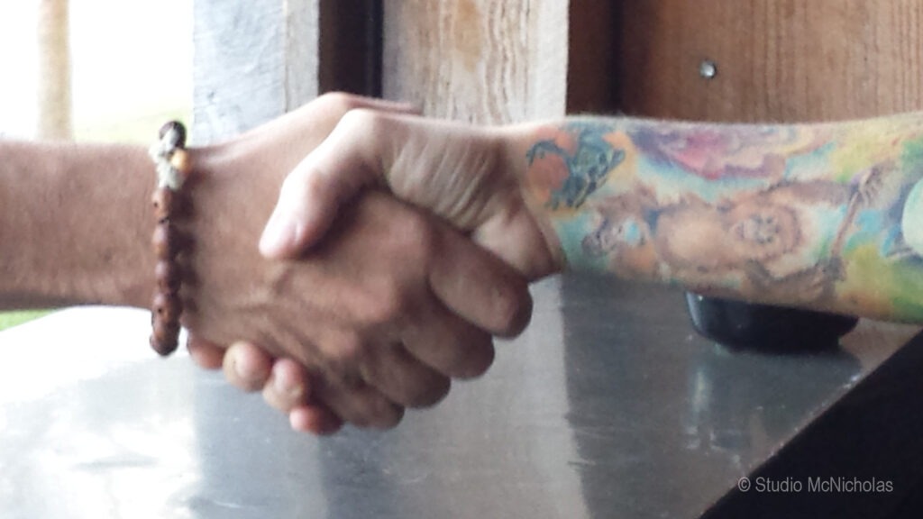 Hands shake in a gesture of agreement, showcasing one bare arm and another adorned with colorful tattoos. The setting suggests a casual environment.