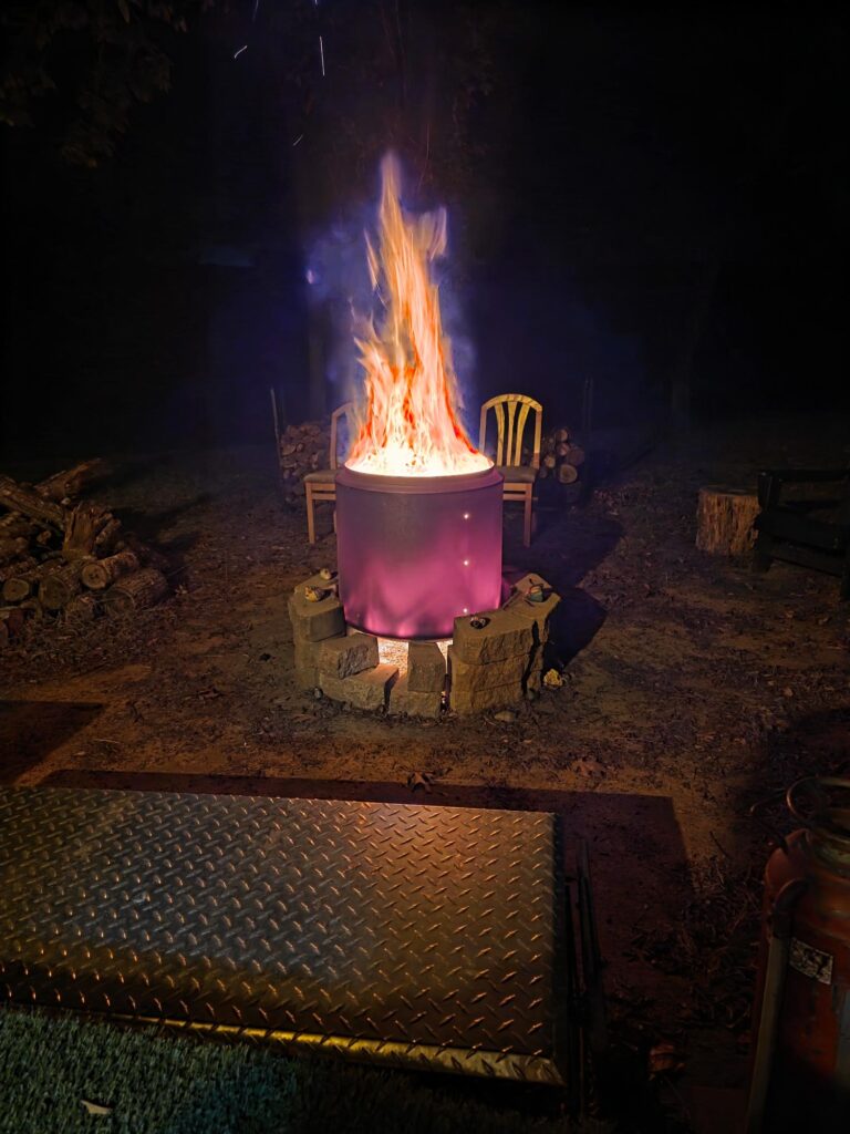 Large purple flames erupt from a metal fire pit at night, surrounded by stone edging and two empty chairs, creating a cozy outdoor atmosphere.