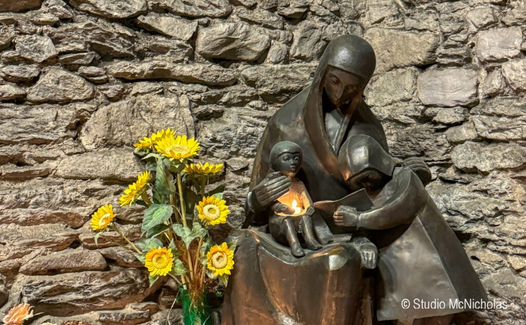 Bronze statue of a mother reading to two children, accompanied by sunflowers, set against a rustic stone wall, symbolizing family and nurturing.