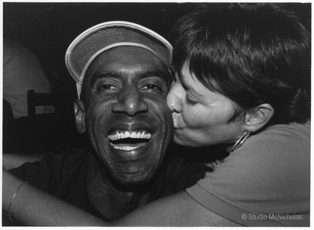 A joyful man smiles widely as a woman kisses his cheek, capturing a moment of affection and camaraderie in a lively setting.