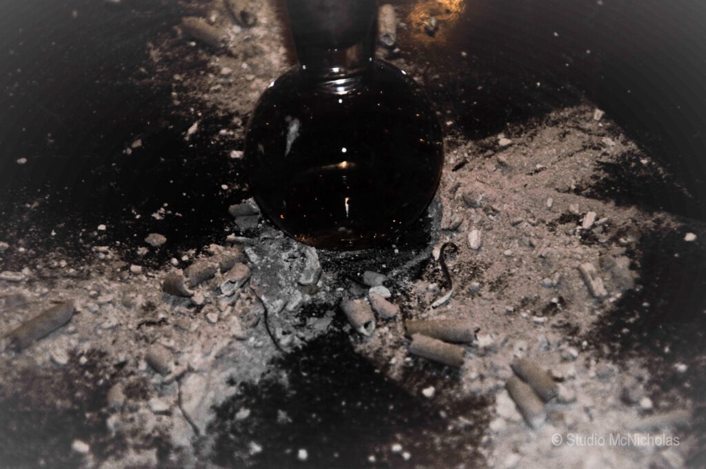 A dark glass vessel sits atop a textured surface scattered with gray ash and debris, suggesting a context of experimentation or ritual.