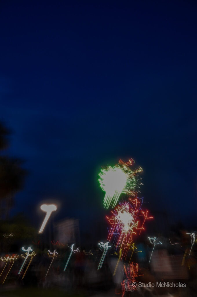Colorful fireworks burst in the night sky, illuminating a blurred crowd and surrounding lights in a festive, vibrant atmosphere.