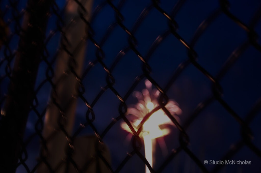 Fireworks burst against a dark sky, their colors blurred by a fence in the foreground, emphasizing the celebration beyond its confines.