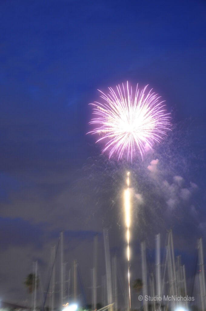 Fireworks burst brightly against a twilight sky, illuminating the silhouette of nearby sailboats in the harbor.