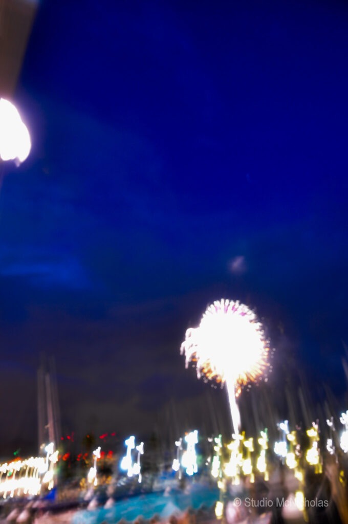 Colorful fireworks burst in a night sky above a harbor, with blurred lights from boats and the shoreline creating a festive atmosphere.
