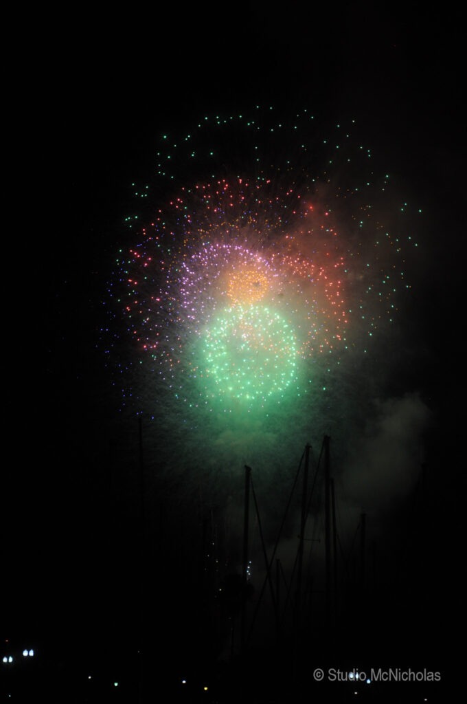 Colorful fireworks burst in the night sky, featuring green, red, and purple hues. Silhouettes of amusement rides are visible below, enhancing the festive atmosphere.