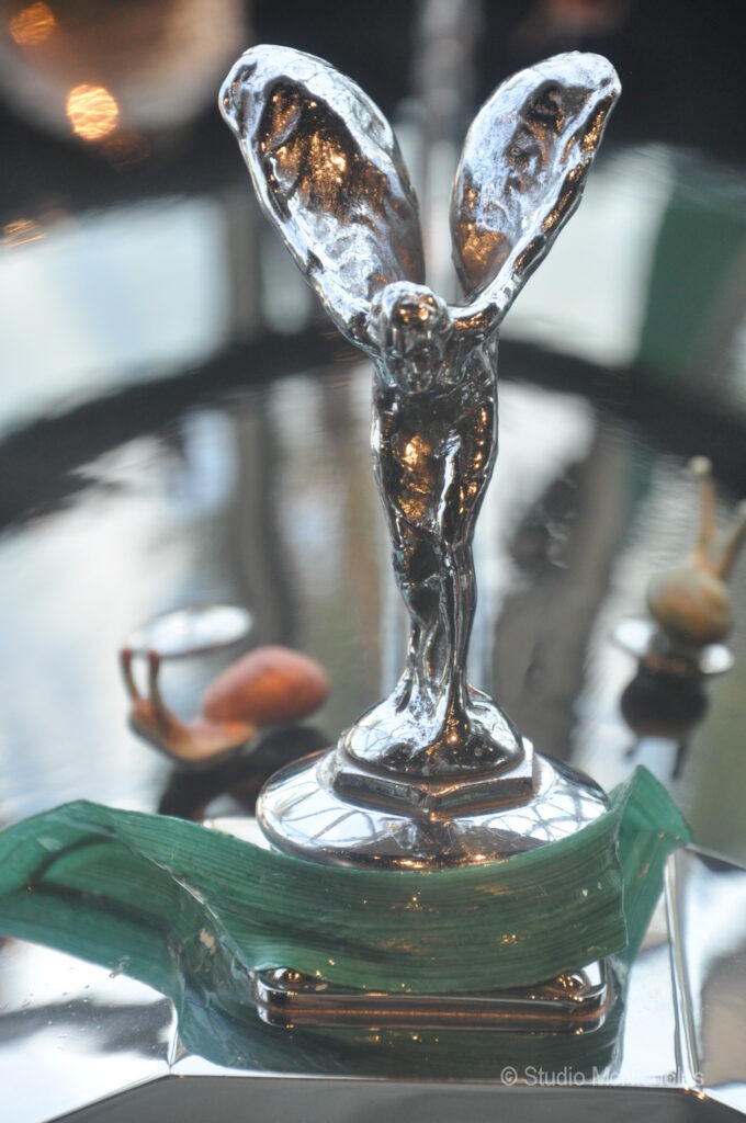 A shiny silver statuette with outstretched wings stands on a reflective surface, surrounded by decorative elements. It symbolizes excellence or achievement.