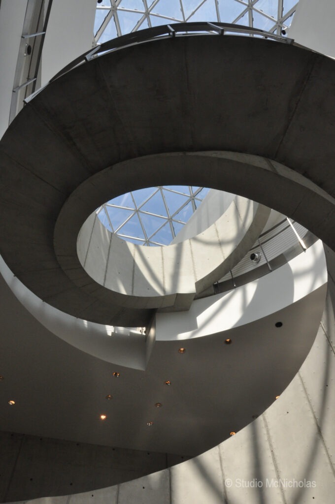 Spiral concrete staircase leading up toward a skylight in a modern architectural space, showcasing light and shadow interplay.