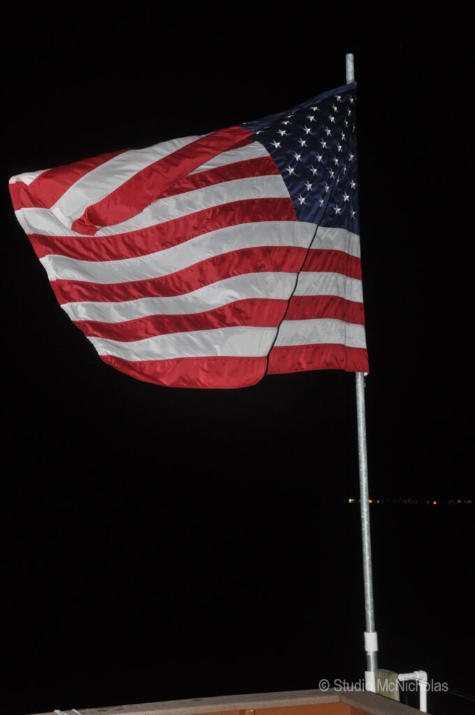 American flag waving prominently against a dark night sky, symbolizing patriotism and unity. The scene captures a moment of national pride.