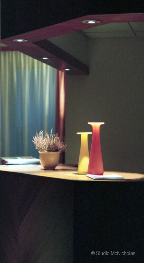 Two vibrant vases, one yellow and one red, alongside a potted plant, are displayed on a wooden table under warm lighting.