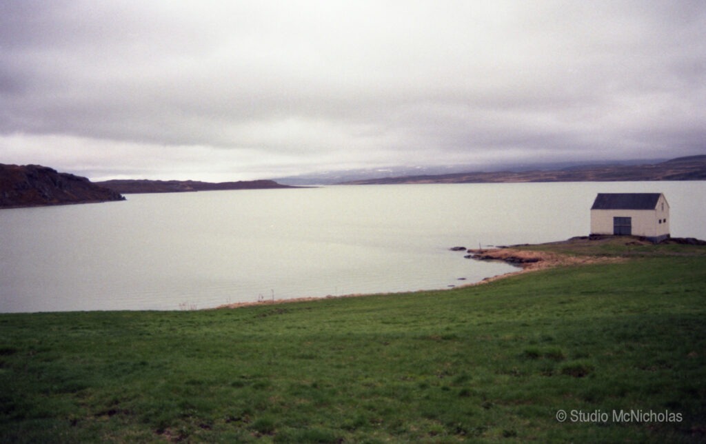 A tranquil view of a lake bordered by green grass, featuring a small white building on the shore under a cloudy sky.