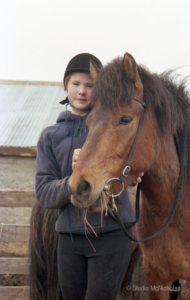 A young rider in a helmet embraces a brown horse at a stable, showcasing the bond between them in a rural setting.