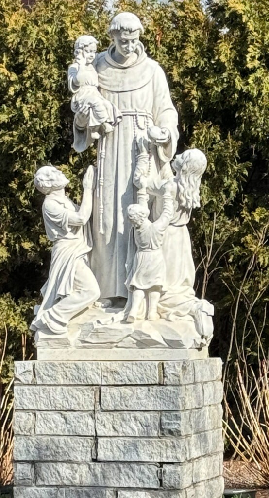 Statue of a saint holding a child, surrounded by two kneeling figures holding children, set against a backdrop of greenery. Symbolizes compassion and care.