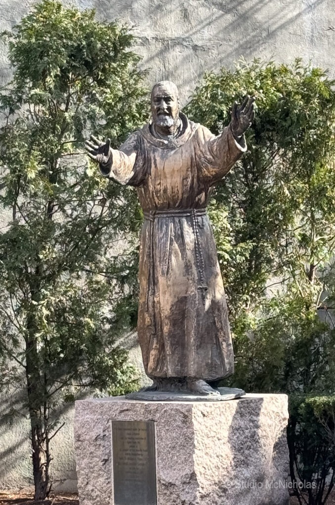 Bronze statue of a historical figure reaching out with open hands, surrounded by greenery, symbolizing peace or guidance.