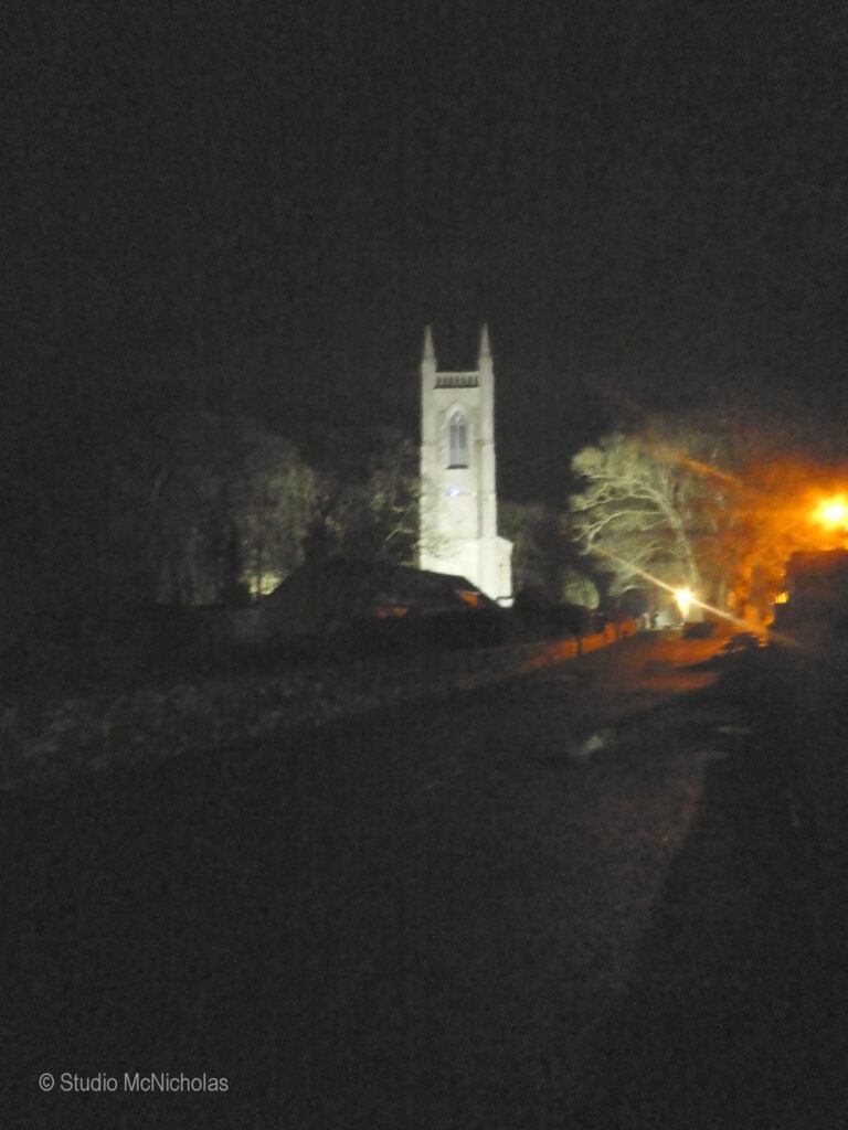 Illuminated church tower at night, set against a dark landscape with faint streetlights and trees in the background.
