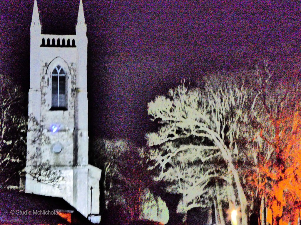 Historic church tower illuminated against a dark night sky, surrounded by bare trees. The haunting atmosphere enhances its architectural details.