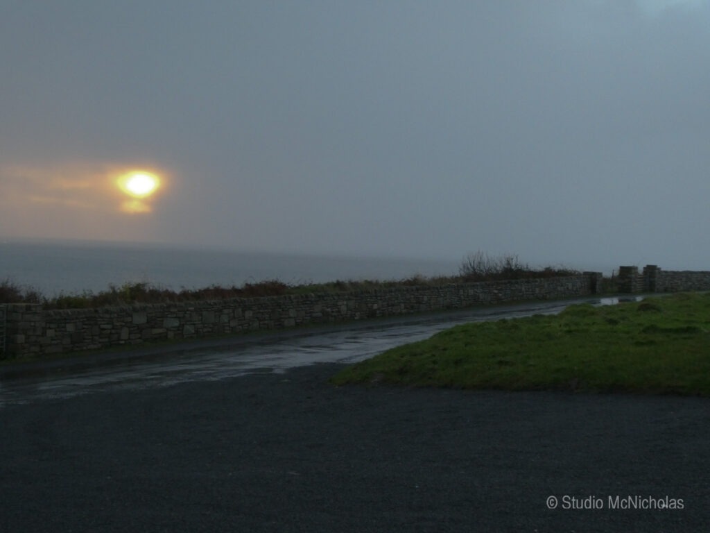 Sun peeking through stormy clouds over the ocean, illuminating a gloomy landscape with a stone wall and wet pavement, suggesting changeable weather.