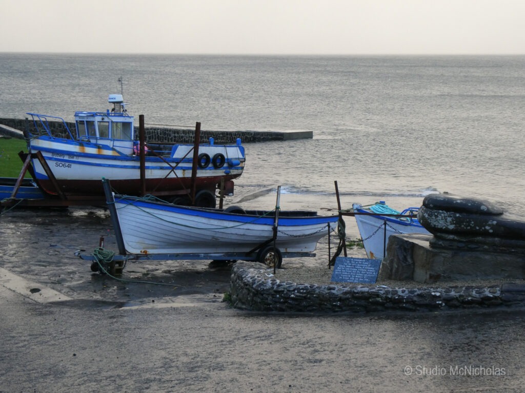 Two fishing boats rest on a sandy shore, surrounded by a rocky wall, with calm sea water in the background. The image highlights a coastal setting.