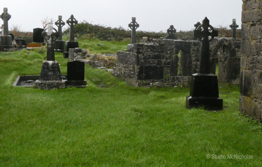 Celtic crosses and gravestones in a grassy cemetery, framed by ancient stone ruins, evoke a somber atmosphere of remembrance.