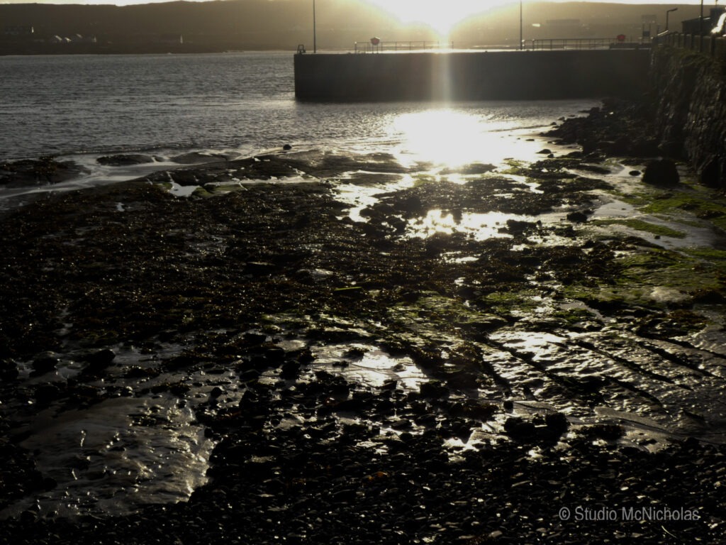 Sunlight reflects off a rocky shore at low tide, revealing wet rocks and seaweed along the coastline, creating a tranquil seaside atmosphere.