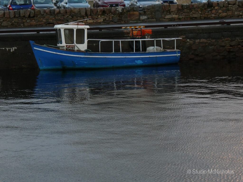 A blue fishing boat is docked in calm waters, reflecting its image. Nearby, parked cars and a stone wall create a serene maritime scene.