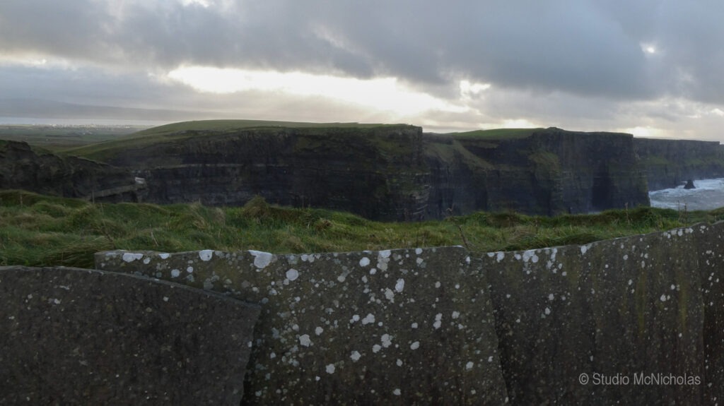 Cliffs rise dramatically above crashing waves, set against a cloudy sky. This captures the rugged beauty of Ireland's coastline at the Cliffs of Moher.