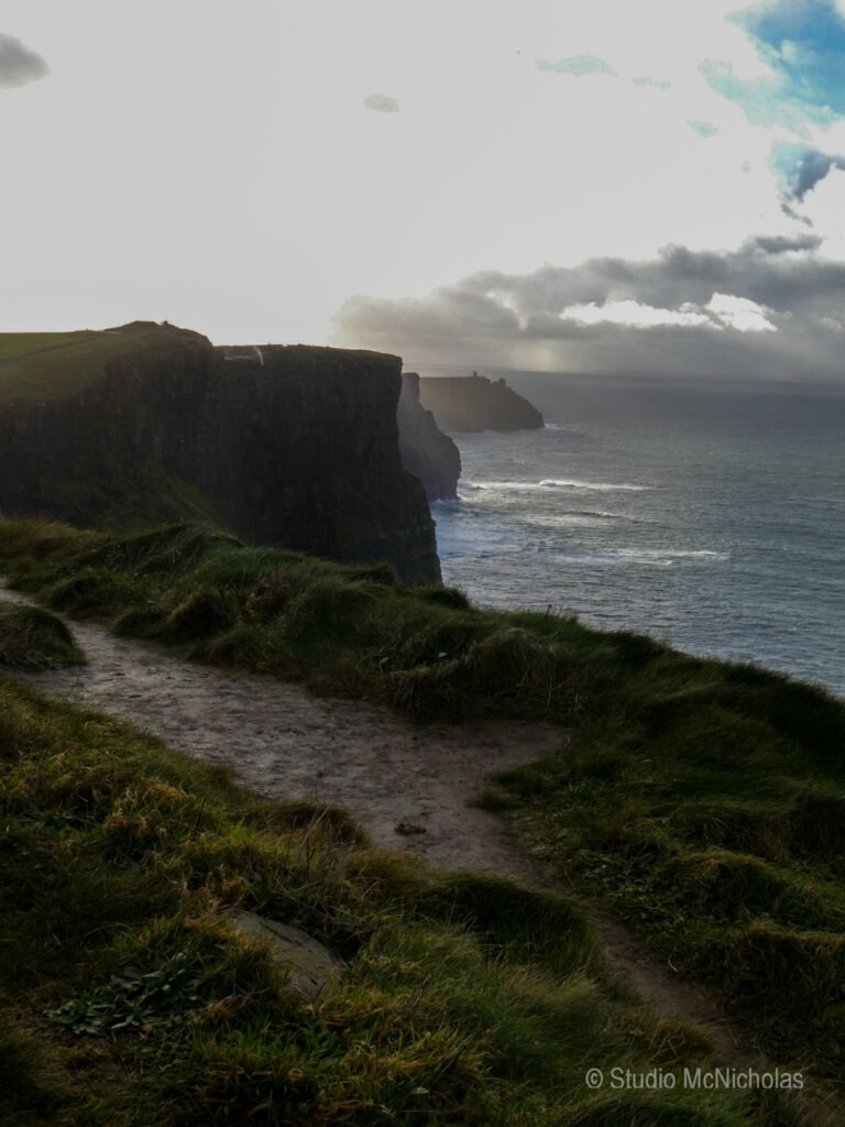 Dramatic cliffs overlooking a turbulent sea, with a lighthouse visible in the distance under a cloudy sky, illustrating coastal beauty and rugged terrain.