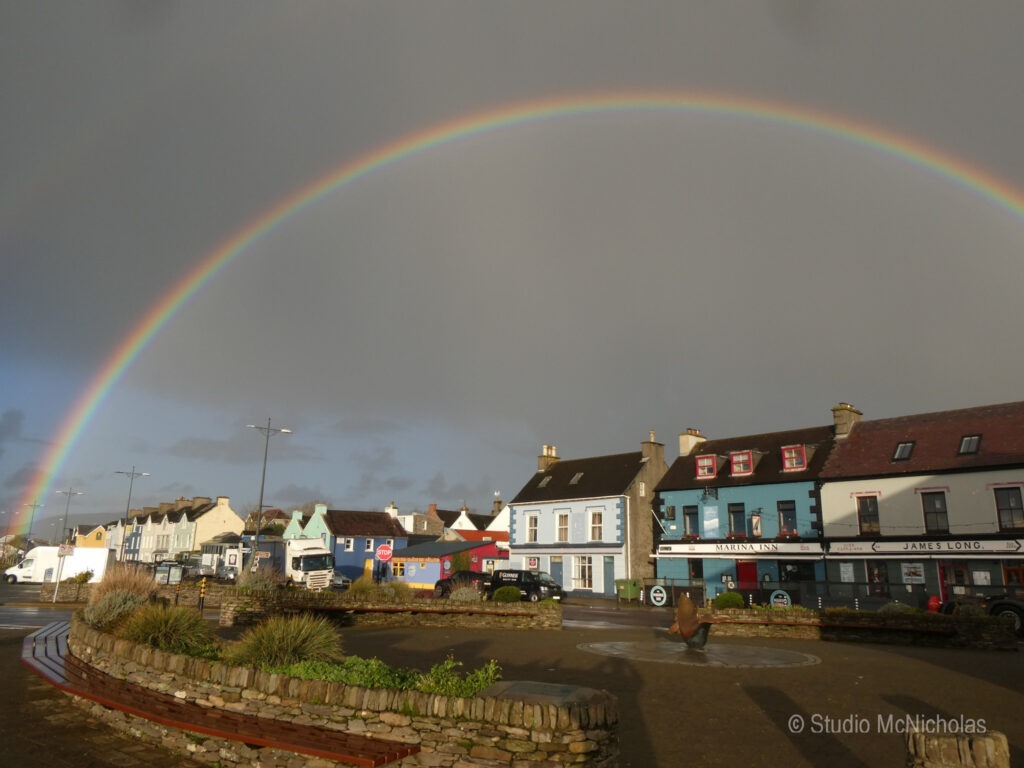 Rainbow arching over a colorful seaside town with shops and a roundabout, highlighting the vibrant atmosphere post-rain.