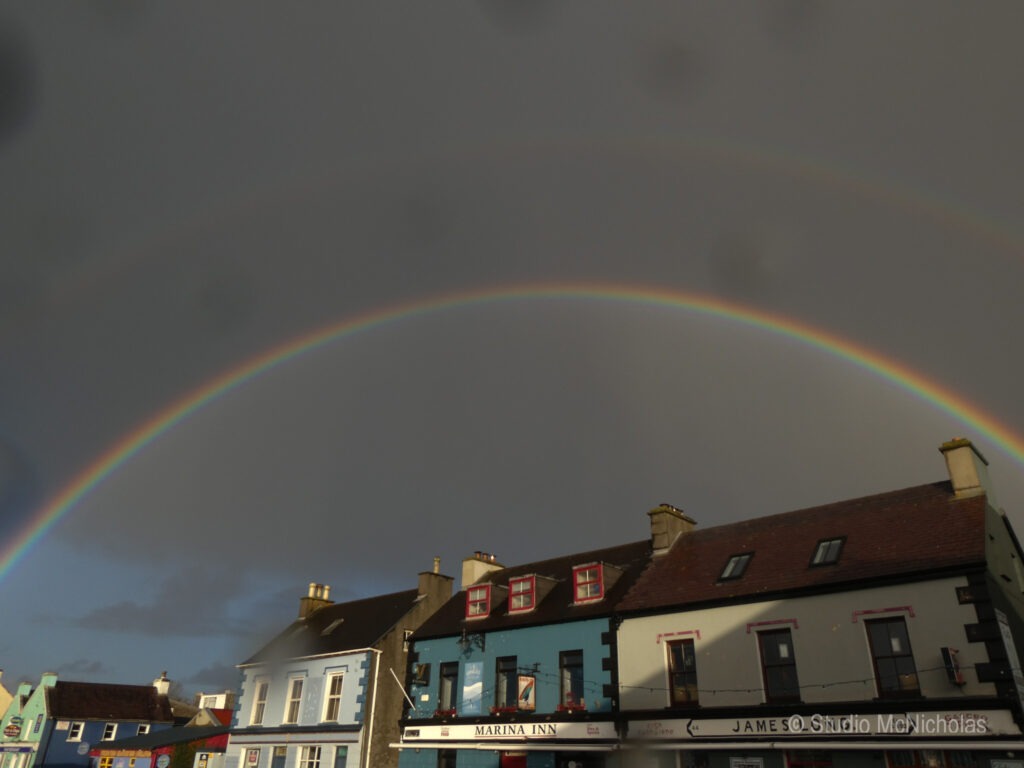 A vibrant double rainbow arches over colorful buildings against a gray sky, highlighting the beauty of nature amidst urban scenery.