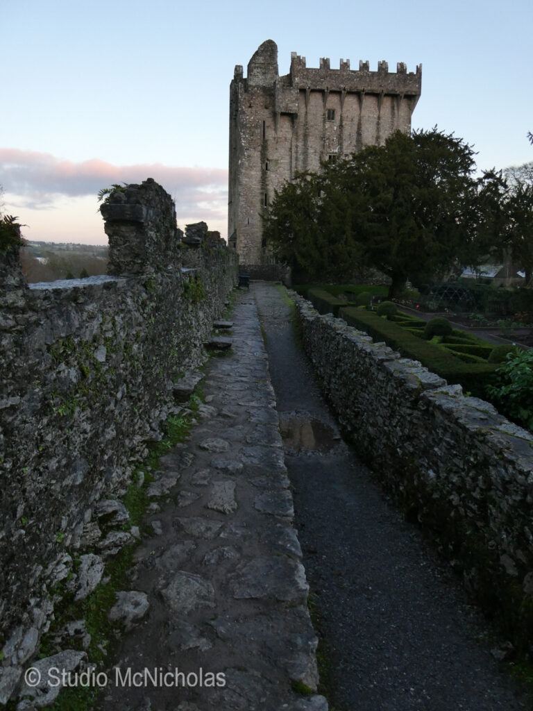 Ballyhannan Castle rises above a stone pathway, surrounded by lush gardens and trees, showcasing Ireland's rich historical architecture.