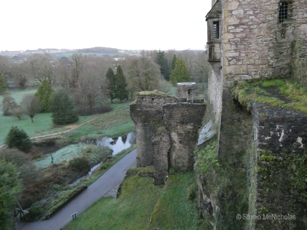Ruins of a stone castle perched on a cliff, overlooking a green landscape with trees and a winding stream below, creating a serene historical scene.