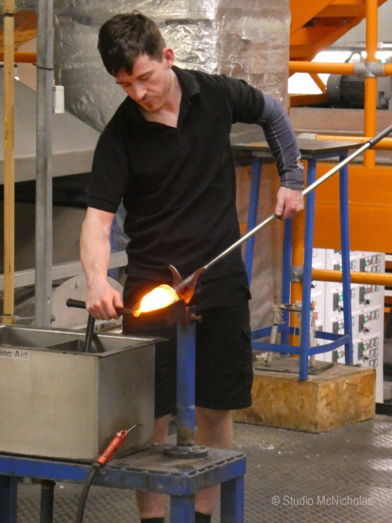 A glassblower shapes molten glass using a metal rod, demonstrating the craftsmanship involved in glass art creation.