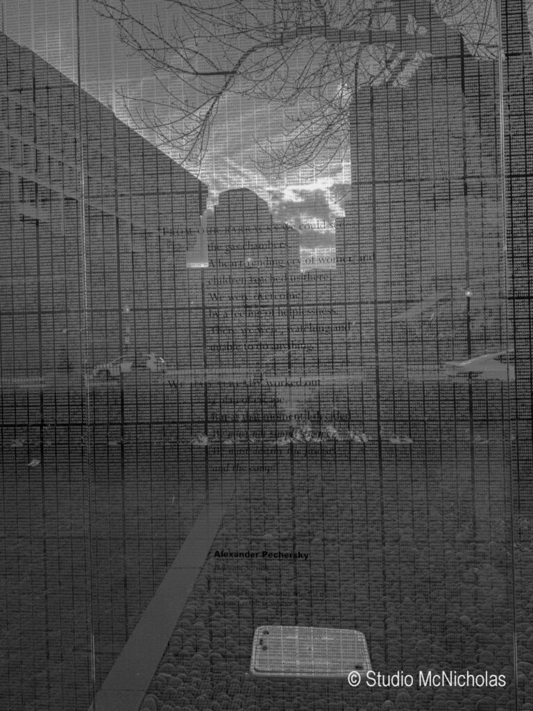 A reflective glass panel displays text about Alexander Pechersky amid a cloudy skyline, symbolizing remembrance and historical narrative.