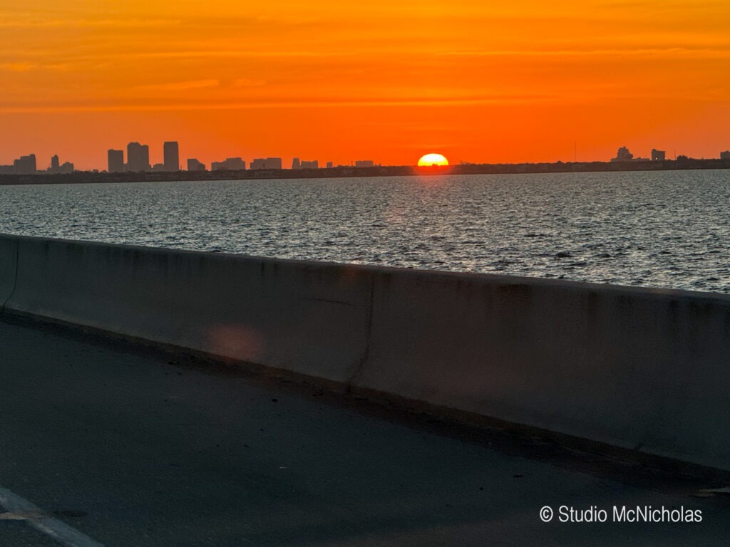 Sunset over a city skyline, with vibrant orange and yellow hues reflecting on the water. A barrier separates the road from the bay.