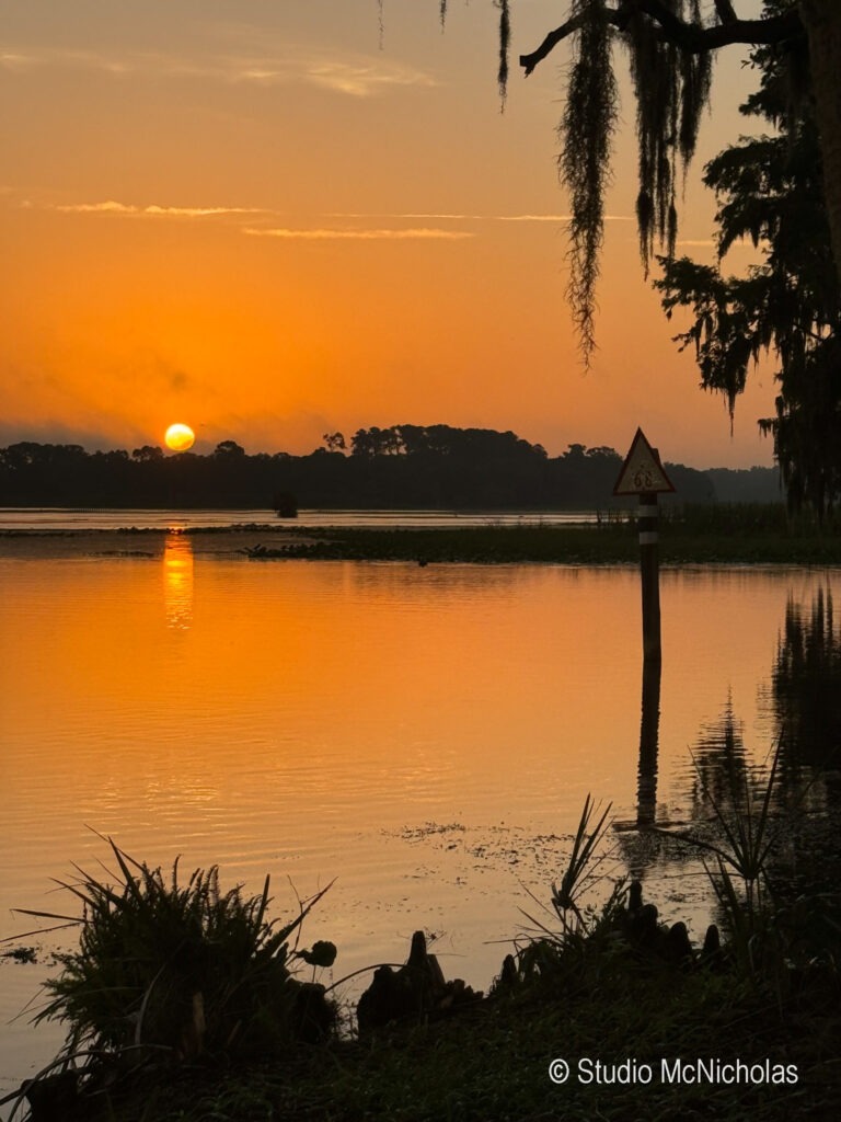 Sunset over a calm lake, with glowing orange hues reflected in the water. Silhouetted trees and a warning sign enhance the tranquil scene.