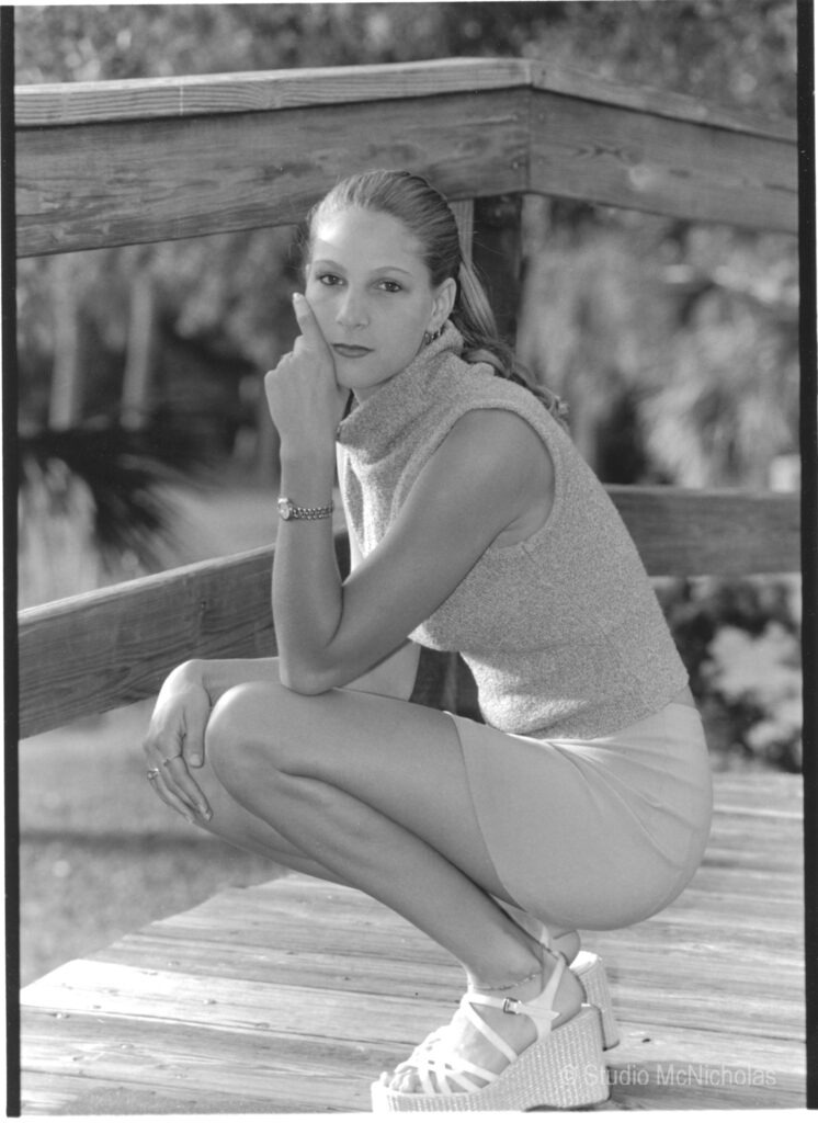 A young woman poses thoughtfully on a wooden deck, wearing a fitted sleeveless top and a short skirt paired with platform sandals.