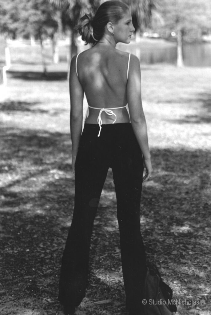 A woman in black pants and a backless white top stands outdoors, gazing back over her shoulder, framed by trees and a lake.