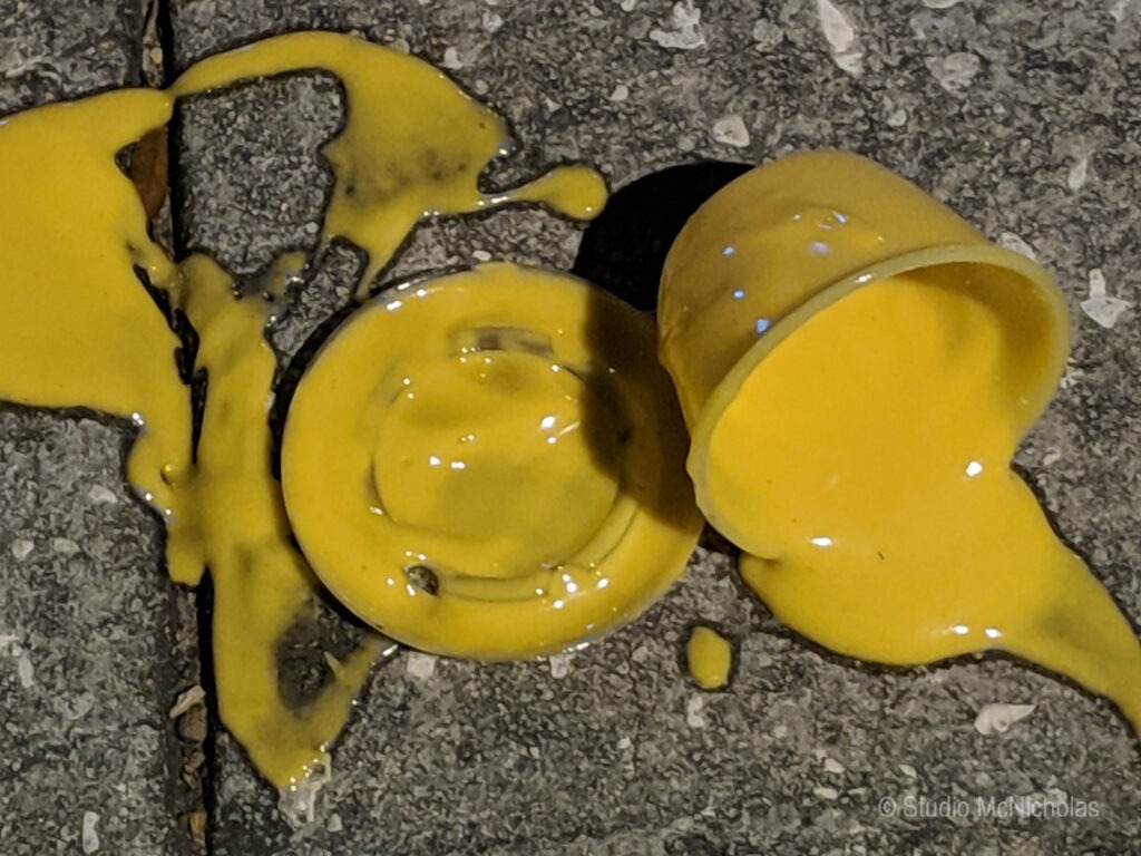 Spilled yellow paint covers a textured gray surface, with a toppled cup and splatters creating a vibrant, messy scene.
