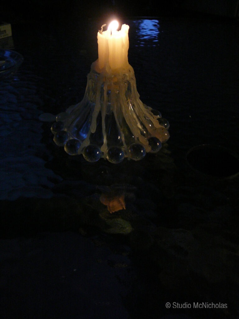 A lit candle drips wax onto a glass holder, reflecting softly on a dark, textured surface, creating a warm, intimate atmosphere.