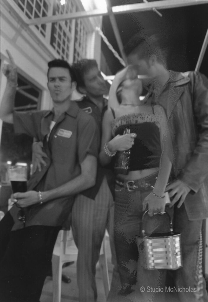 Four friends pose playfully at a party, with two men and a woman interacting closely. The scene is lively, capturing a joyful social moment.