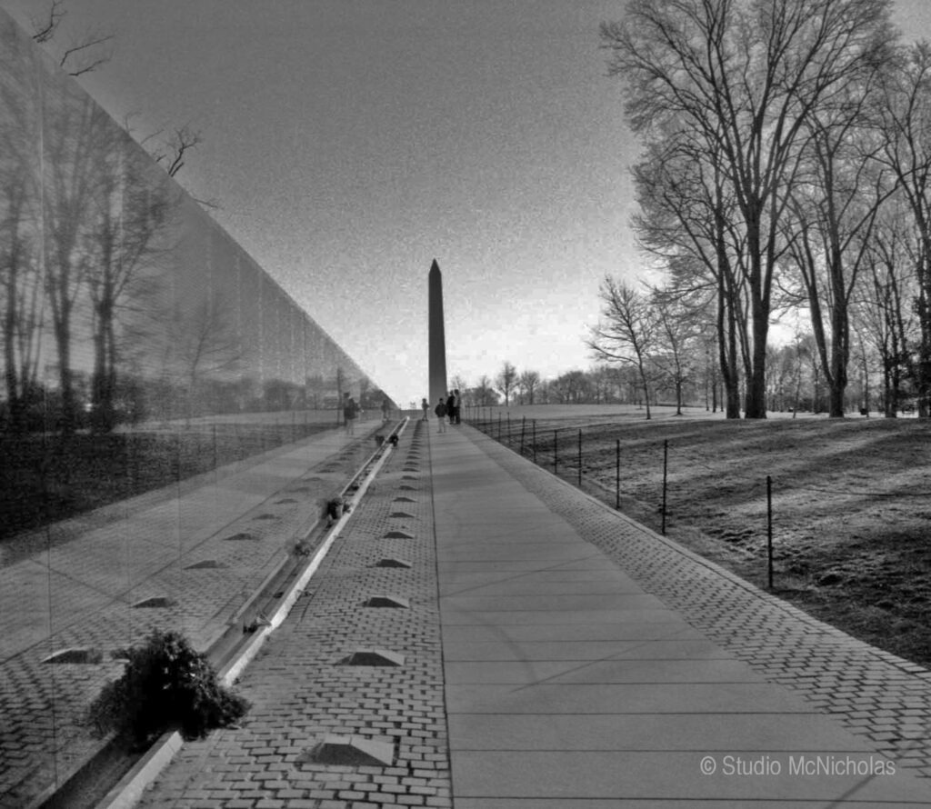 Vietnam Veterans Memorial wall reflects the Washington Monument, surrounded by trees. People pay their respects along the walkway.