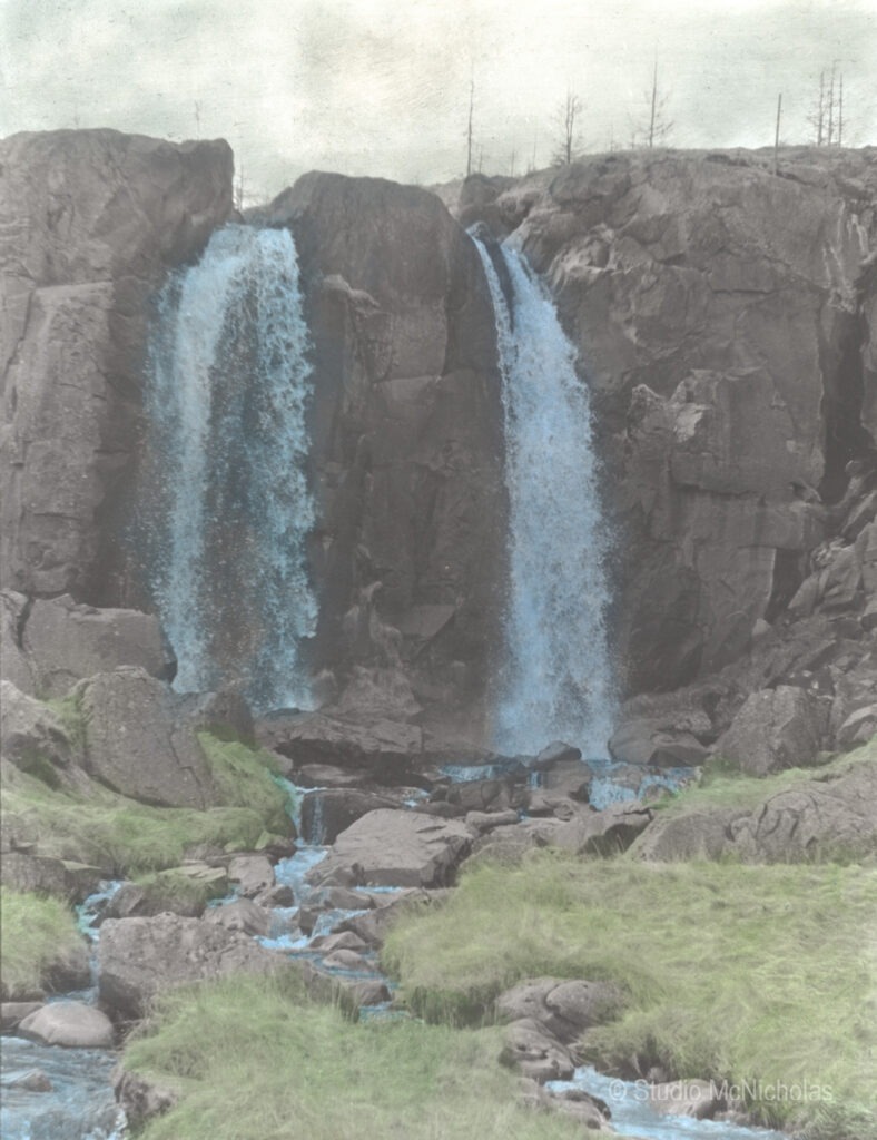 Two waterfalls cascade over rocky cliffs, surrounded by greenery and flowing water. The scene captures the beauty of nature's landscape.
