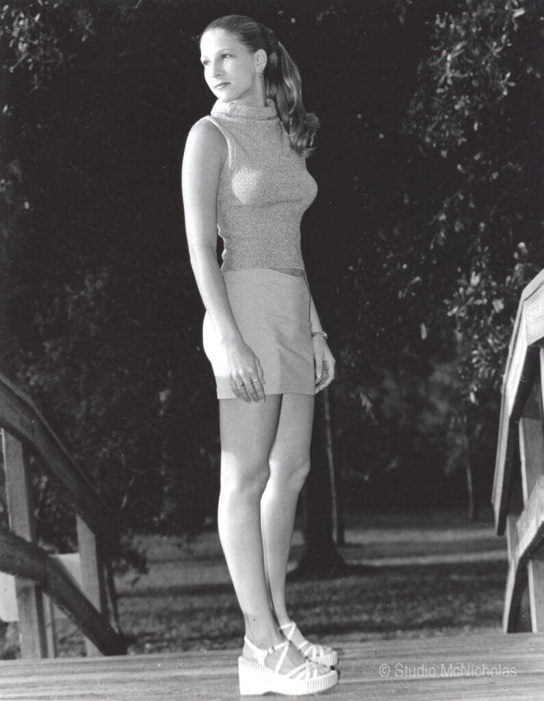 Young woman in a fitted sleeveless top and short skirt stands confidently on wooden steps, surrounded by greenery. The black-and-white photo captures a chic, casual moment.